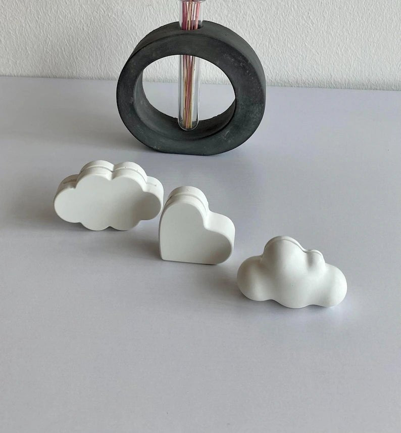 Three white cloud and heart-shaped figurines, with a round grey vase holding colourful dry flowers in the background.