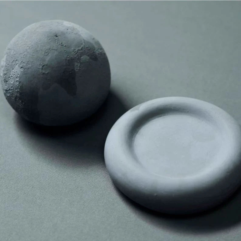 A textured grey sphere resembling a moon next to a smooth, round grey dish on a muted background.