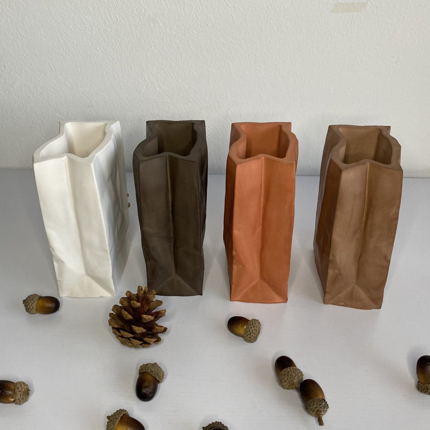 Four paper bag-style ceramic vases in white, dark brown, terracotta, and bronze, with acorns and a pinecone on the table.