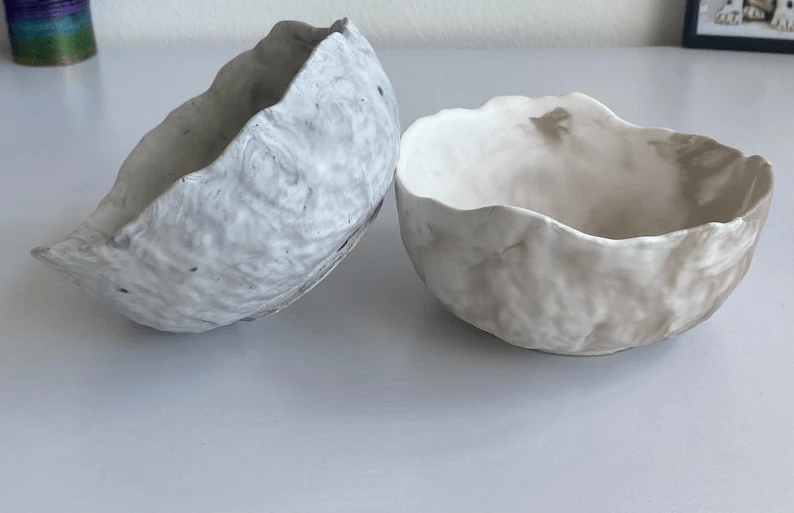 Two textured ceramic bowls, one light grey and the other beige, stacked on a white surface with a minimalist background.