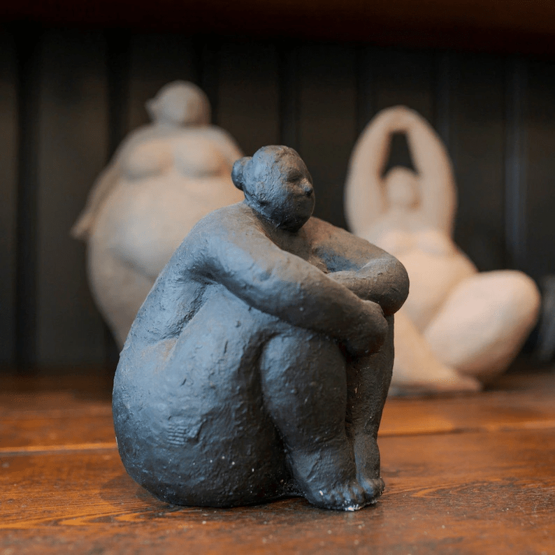 A small, seated black sculpture with rounded features, surrounded by other abstract figures in a warm-toned wooden setting.