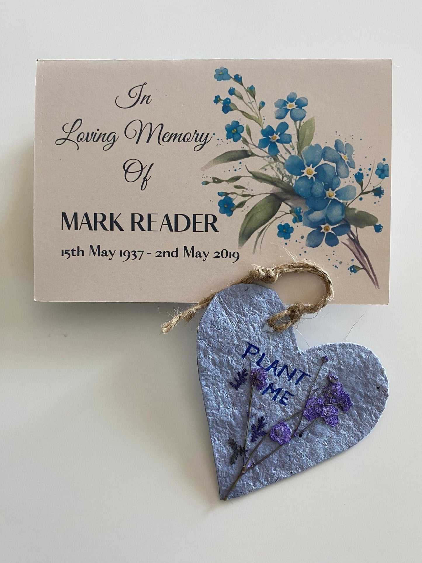 A memorial card featuring a blue floral design, a heart-shaped plantable tag saying "PLANT ME," and a decorative glass bear.