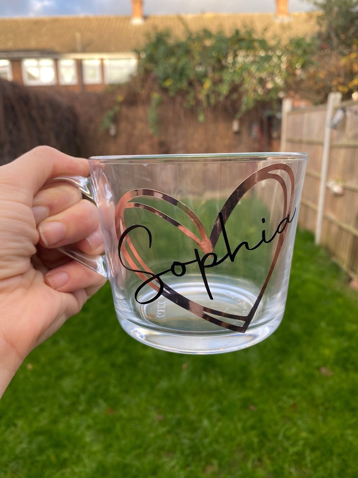 Hand holding a clear mug with a heart design and the name "Sophia" printed in elegant black against a green lawn backdrop.