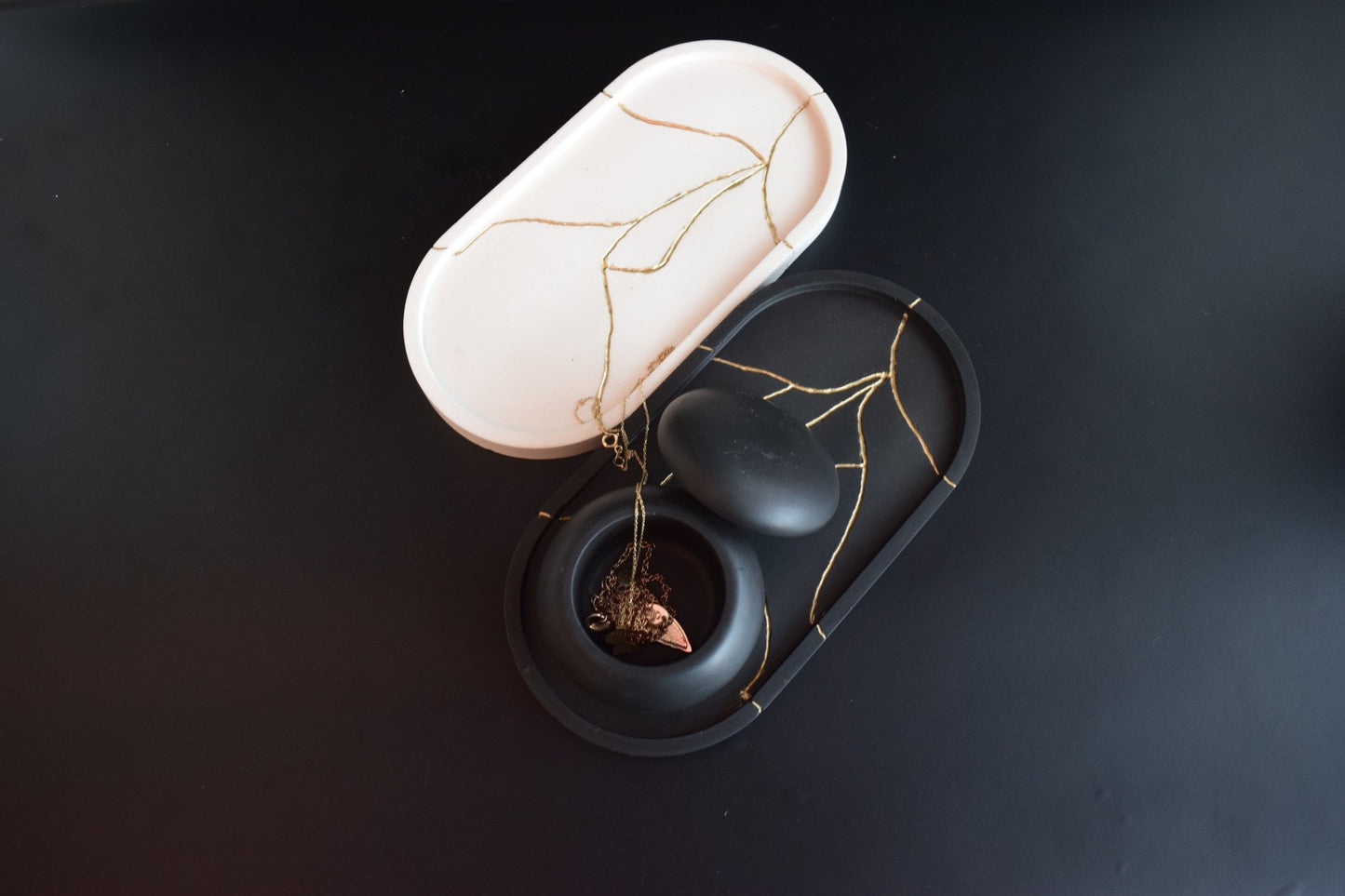 Black and white oval trays with gold cracks on a dark surface. Black tray holds a small black bowl with a tangled necklace.