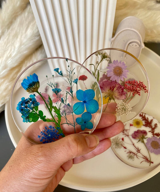 A hand holds two round resin coasters featuring colourful pressed flowers against a soft background with textured decor.