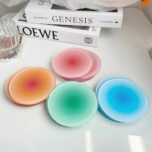 Four colourful gradient coasters in peach, pink, green, and blue lie on a white table, alongside stacked books and a glass.