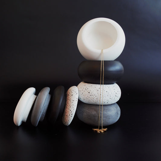 A stack of oval stone pots in white to black, with lids, against a black background, a delicate gold necklace draped over